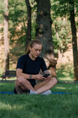 Spor minderinde, elinde akıllı bir telefonla parkta dinlenen genç bir bayan sporcunun portresi. Sağlıklı yaşam tarzı kavramı.