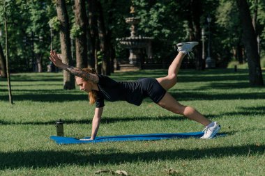 Genç sporcu kız spor paspası üzerinde egzersiz yapıyor. Parktaki çimenlerde bacaklarını sallıyor. Sağlıklı yaşam tarzı kavramı.