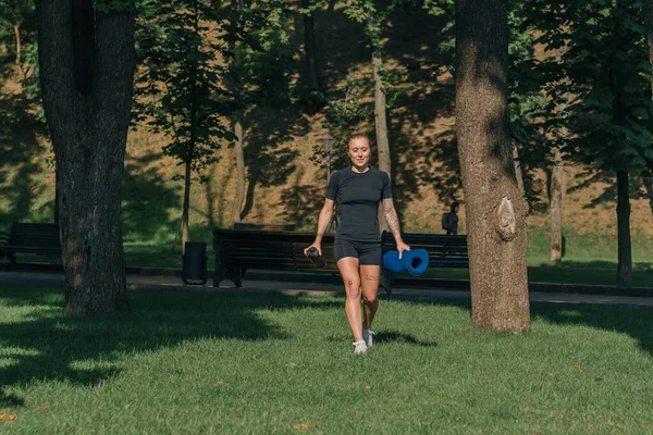 年轻的女运动员在公园外训练前 手里拿着皮垫和瓶装水 把草垫放在健康的生活方式概念上 — 图库照片