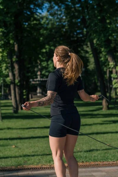 ヘルスパークの街で運動中にスポーツジャンプロープをしている朝のタトゥーを持つフィットネス若いスポーツマンガール — ストック写真
