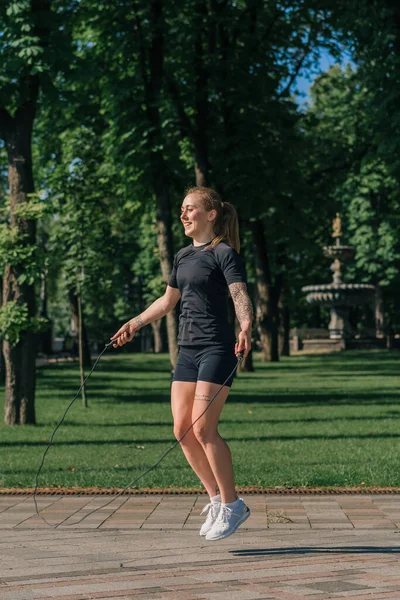 ヘルスパークの街で運動中にスポーツジャンプロープをしている朝のタトゥーを持つフィットネス若いスポーツマンガール — ストック写真