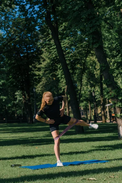 年轻的女运动员在运动垫上做体育锻炼 腿上绑着橡皮筋 进行公园外健康生活方式的概念 — 图库照片