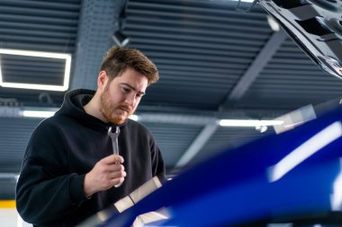Elinde İngiliz anahtarı olan bir adam, bozuk mavi arabasının açık kaputunda duruyor ve nasıl tamir edileceğini düşünüyor.