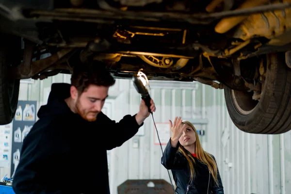 A girl shows a car mechanic with a flashlight what she has broken in the suspension of her luxury car which is standing on a lift in car service