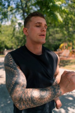 Şehir parkında spor kıyafetleriyle koşan, dövmeli, genç, bitkin bir koşucu.