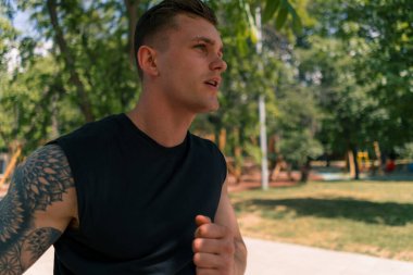 Şehir parkında spor kıyafetleriyle koşan, dövmeli, genç, bitkin bir koşucu.