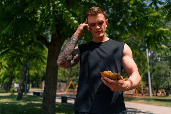 若いスポーツマンは 彼の手で甘いドーナツを保持トレーニングした後 ジャンクフードを食べるかどうか躊躇します — ストック写真