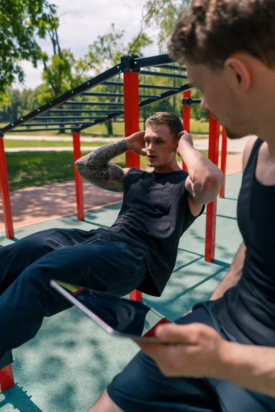 a fitness trainer with a tablet in his hands trains a young athlete pumping his abs on the street on sports ground motivation support