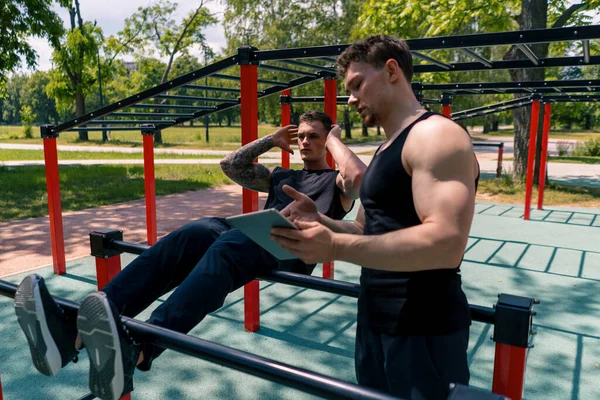 a fitness trainer with a tablet in his hands trains a young athlete pumping his abs on the street on sports ground motivation support