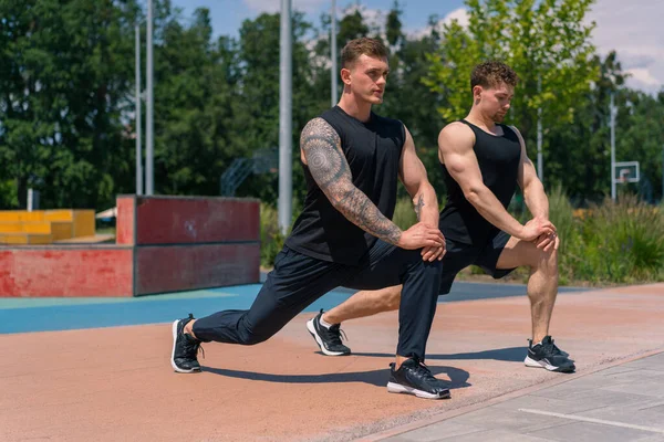 Personal fitness trainer training athlete on sports field Two active sweaty guys stretching leg outdoors doing exercises