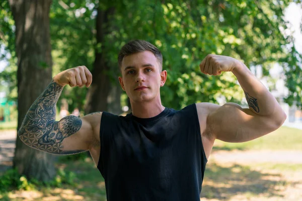 portrait of young slim sportsman with tattoos and piercings showing pumped biceps outdoors street workout park motivation