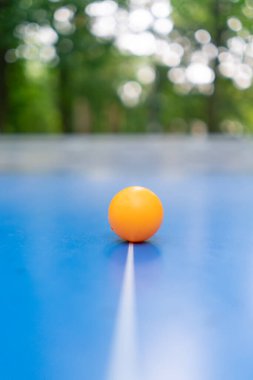 Turuncu bir tenis topu, şehir parkındaki bir ağın yanındaki mavi tenis masasına dayanır. Açık havada pinpon oynama kavramı.