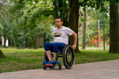 Özürlü bir adam, şehir parkı yolunda, arka plandaki ağaçlara karşı tekerlekli sandalyeye biner.