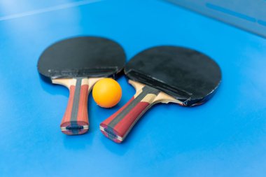 İki tenis raketi ve turuncu bir tenis topu, şehir parkındaki masa tenisi oyununun yakınındaki ağın yanında, mavi bir tenis masasının üzerinde yatıyor.