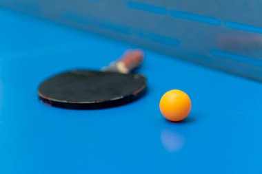 Tenis raketi ve turuncu tenis topu, şehir parkındaki masa tenisi oyununun yakınındaki ağın yanındaki mavi tenis masasının üzerinde.