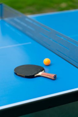 Tenis raketi ve turuncu tenis topu, şehir parkındaki masa tenisi oyununun yakınındaki ağın yanındaki mavi tenis masasının üzerinde.
