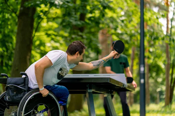 Tekerlekli sandalyedeki engelli bir adam, şehir parkında yaşlı bir adamla ağaç zeminine karşı pinpon oynuyor.