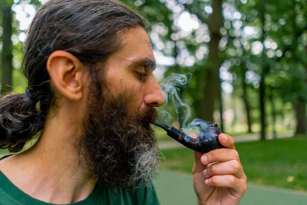 長い髪と灰色のひげを持つ老人の肖像画 木の背景に対する市立公園のパイプを喫煙 — ストック写真