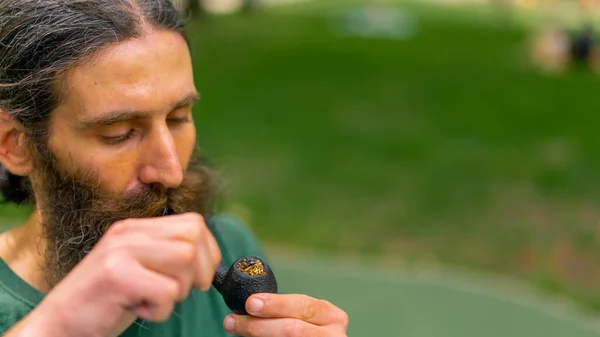 長い髪と灰色のひげを持つ老人の肖像画 木の背景に対する市立公園のパイプを喫煙 — ストック写真