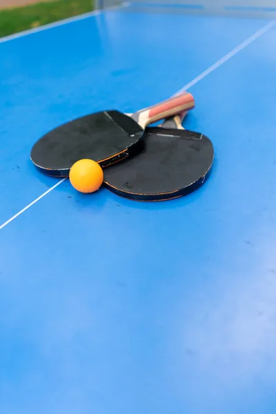 Two tennis rackets and an orange tennis ball lie on a blue tennis table next to a net in a city park ping pong game