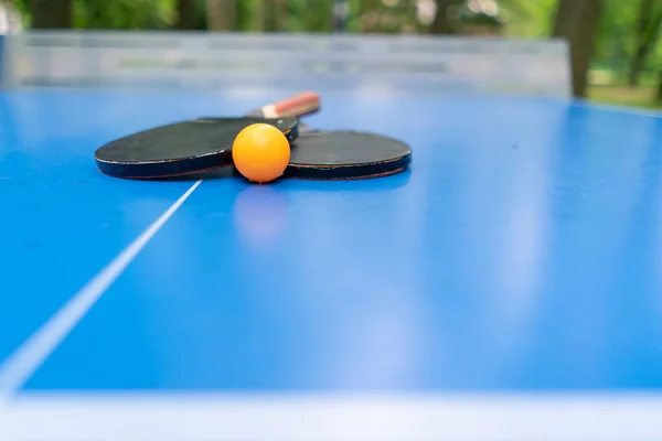 2つのテニスラケットとオレンジテニスボールは 都市公園のピンポンゲームのネットの隣にある青いテニステーブルの上にあります — ストック写真