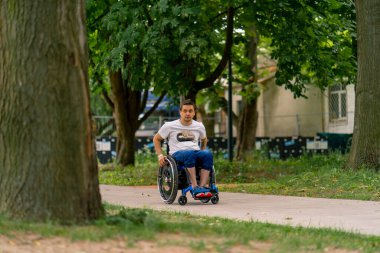 Engelli bir gencin özgünlük portresi. Bir şehir parkında tekerlekli sandalyede, ağaçların arkasında.