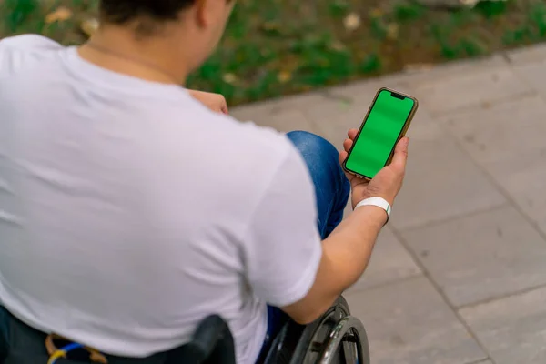 内包性 車椅子に障害のある男性が後ろから市立公園の景色を眺めるオープングリーンスクリーン付きの携帯電話を持っています — ストック写真