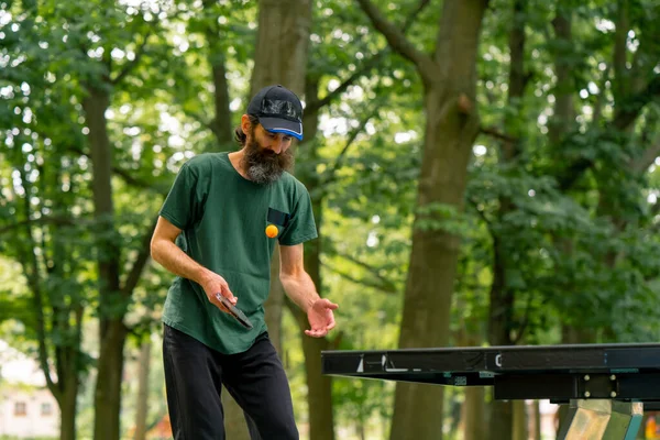 キャップを身に着け ラケットを持っている年上の男性は 木の背景に対して市の公園でピンポンを演奏します — ストック写真