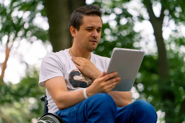 包容性 車椅子に障害のある男性は 彼が公園で保持しているタブレットを意図的に見ています — ストック写真
