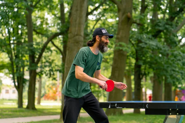 キャップを身に着け ラケットを持っている年上の男性は 木の背景に対して市の公園でピンポンを演奏します — ストック写真