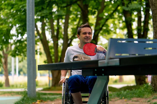 一个坐轮椅的残疾人在城市公园的树后打乒乓球 — 图库照片