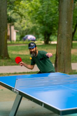 Uzun saçlı ve gri sakallı yaşlı bir adam şehir parkında masa tenisi oynuyor.