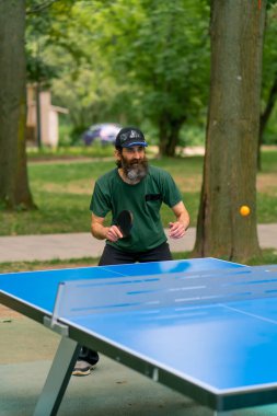Uzun saçlı ve gri sakallı yaşlı bir adam şehir parkında masa tenisi oynuyor.