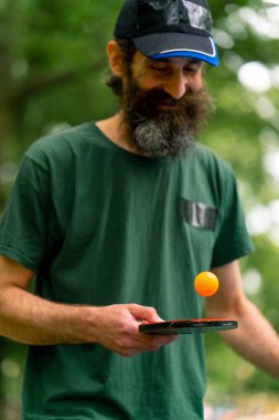 Mavi pinpon masasının yanındaki yaşlı bir adam şehir parkında tenis raketinde turuncu bir topa vuruyor.