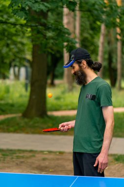 Mavi pinpon masasının yanındaki yaşlı bir adam şehir parkında tenis raketinde turuncu bir topa vuruyor.