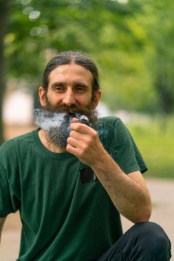 Uzun saçlı ve gri sakallı yaşlı bir adam şehir parkında arka planda ağaçlarla pipo içiyor.