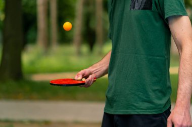 Mavi pinpon masasının yanındaki yaşlı bir adam şehir parkında tenis raketinde turuncu bir topa vuruyor.