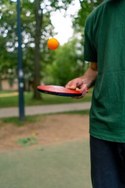 Mavi pinpon masasının yanındaki yaşlı bir adam şehir parkında tenis raketinde turuncu bir topa vuruyor.