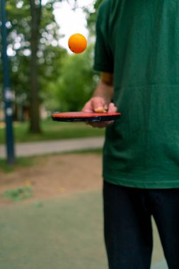 Mavi pinpon masasının yanındaki yaşlı bir adam şehir parkında tenis raketinde turuncu bir topa vuruyor.