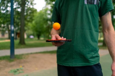 Mavi pinpon masasının yanındaki yaşlı bir adam şehir parkında tenis raketinde turuncu bir topa vuruyor.