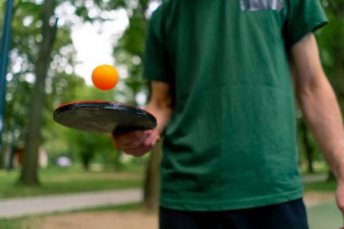 Mavi pinpon masasının yanındaki yaşlı bir adam şehir parkında tenis raketinde turuncu bir topa vuruyor.