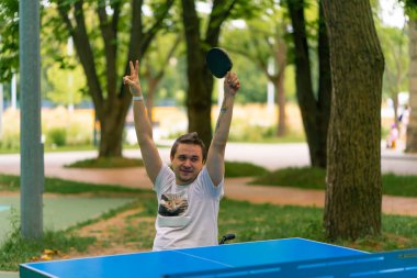 Elinde raketi olan engelli bir adam şehir parkının arka planında pinpon oyunu kazanmasını kutluyor.