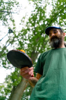 Mavi pinpon masasının yanındaki yaşlı bir adam şehir parkında tenis raketinde turuncu bir topa vuruyor.