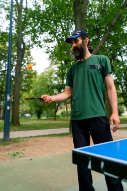 Mavi pinpon masasının yanındaki yaşlı bir adam şehir parkında tenis raketinde turuncu bir topa vuruyor.
