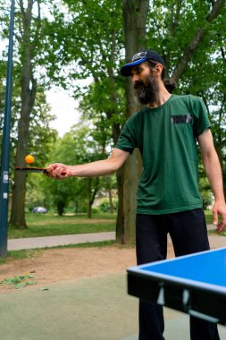 Mavi pinpon masasının yanındaki yaşlı bir adam şehir parkında tenis raketinde turuncu bir topa vuruyor.