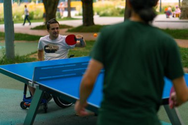 Mutlu Tekerlekli sandalyedeki engelli adam şehir parkında sakallı yaşlı bir adama karşı ping pong oynuyor.