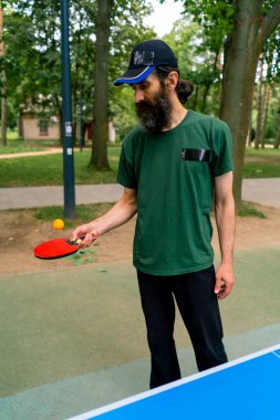 Mavi pinpon masasının yanındaki yaşlı bir adam şehir parkında tenis raketinde turuncu bir topa vuruyor.