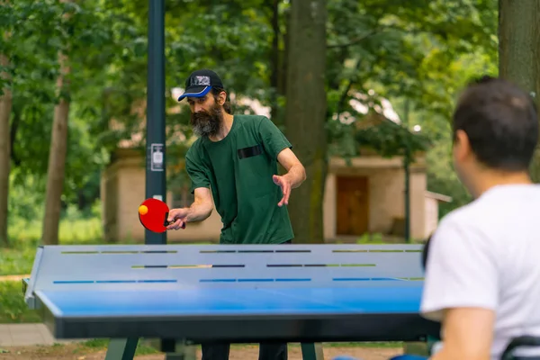 一个坐轮椅的残疾人在城市公园里和一个留着灰胡子的老人打乒乓球 — 图库照片