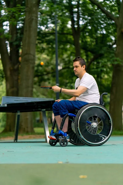 在城市公园的一个特写镜头里 坐在轮椅上 坐在蓝色乒乓球桌旁的包容性残疾人在网球拍上打了一个橙色球 — 图库照片