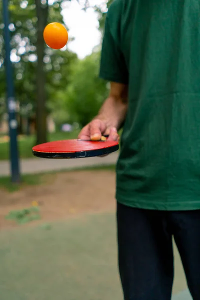 一位老人坐在蓝色乒乓球桌旁 在一个城市公园的网球拍上打了一个橙色球 — 图库照片